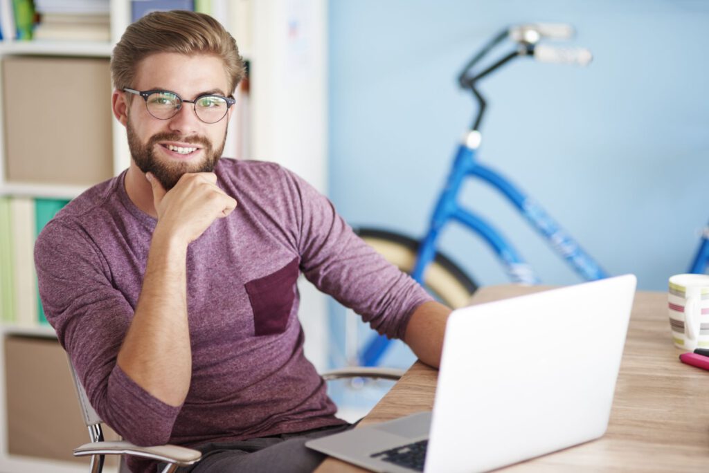 Freundlicher junger Mann mit Brille und Bart am Schreibtisch mit Laptop, im Hintergrund ein Fahrrad